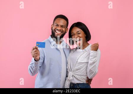 Aufgeregt schwarzes Paar mit Kreditkarte, die ja Geste macht, Förderung kontaktloses Einkaufen über rosa Studio Hintergrund Stockfoto