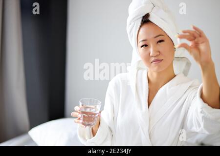 asiatische Frau, die Medizin nach dem Aufwachen in den Morgen, Frau im Bademantel und Handtuch haben Medikamente Schmerzmittel leiden an morgendlicher Krankheit oder Heada Stockfoto