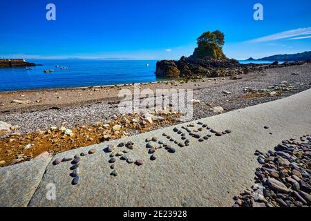 Bild von einem Kiesstrand in der frühen Morgensonne Mit einer kleinen Insel und Hafen im Hintergrund mit Die Wörter REISEN geschrieben in Kieselsteinen in Th Stockfoto