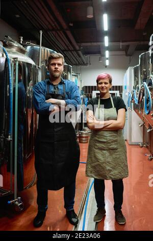 Junge männliche und weibliche Arbeiter der Bierfabrik in Schürzen, die Arme an der Brust kreuzen, während sie vor stehen Kamera Stockfoto