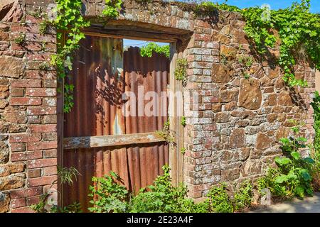 Bild einer alten Steinmauer mit einer alten Türöffnung mit Ziegelumrandung, die im Sonnenschein mit rostigen, korigierten Eisenblechen versperrt wurde. Stockfoto