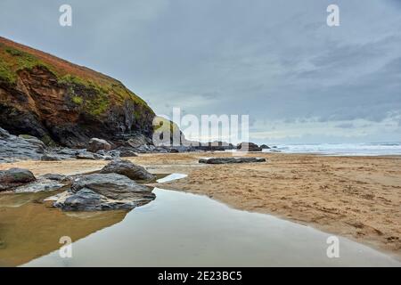 Bild von Poldhu Beach in Cornwall mit Felsen, Wellen, Sand, Wasser im Vordergrund und bewölktem Himmel. Stockfoto