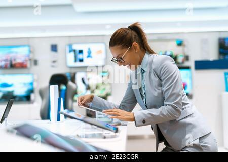 Glückliche kaukasische Frau mit Pferdeschwanz in Business-Kleidung gekleidet suchen fort neue Tablette, indem sie in der Nähe stehen Stand in Tech-Shop. Stockfoto