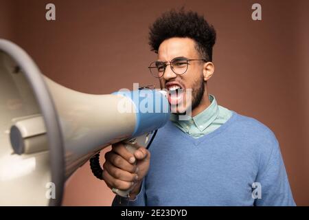 Sehr irritiert junger Mixed-Race-Geschäftsmann oder Chef in Brillen und casualwear schreit laut im Megaphon vor der Kamera Stockfoto