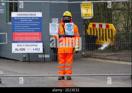 Amersham, Buckinghamshire, Großbritannien. Januar 2021. Ein HS2-Wachmann steht neben einem Coronavirus-Schild am Ortseingang. Trotz der Zahl der Covid-19-Fälle, die außer Kontrolle geraten, dürfen die HS2-Bauarbeiter immer noch durch die neueste Sperre arbeiten. Es wird gefordert, dass die Bauarbeiten im Vereinigten Königreich aufgrund des Risikos einer Übertragung der Covid-19-Krankheit unverzüglich eingestellt werden. Die irische Regierung hat alle nicht notwendigen Bauarbeiten in Irland eingestellt. Quelle: Maureen McLean/Alamy Live News Stockfoto