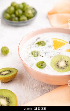 Joghurt mit Kiwi, Stachelbeere, Chia in Keramikschale auf grauem Betongrund und orangefarbenem Leinentextilien. Seitenansicht, Nahaufnahme, selektiver Fokus. Stockfoto