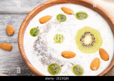 Joghurt mit Kiwi, Stachelbeere, Chia und Mandeln in Holzschüssel auf grauem Holzhintergrund und Leinentextilien. Seitenansicht, Nahaufnahme, selektiver Fokus. Stockfoto