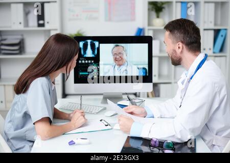 Zwei junge Praktizierende in Uniform sitzen vor dem Schreibtisch Von Computer-Monitor und Blick auf Senior Arzt während Online Kommunikation Stockfoto