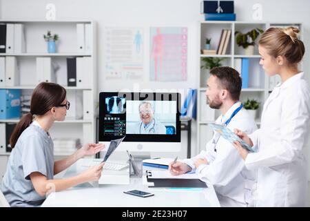 Gruppe junger Praktizierender, die vor dem Schreibtisch sitzen Computermonitor und Beratung mit dem Oberarzt während der Online-Kommunikation Stockfoto