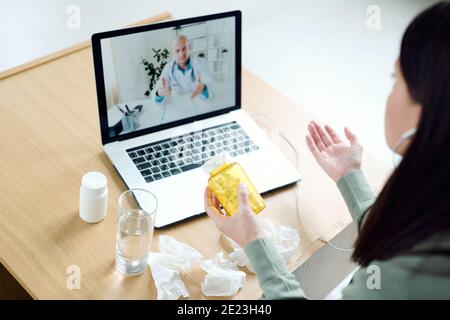 Reife männliche Arzt auf Laptop-Display geben medizinische Empfehlungen zu Junge kranke Patientin mit einer Flasche Pillen, die sie beschreibt Symptome Stockfoto