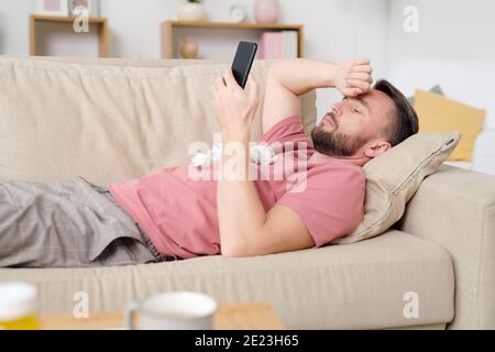Junge kranke männliche Patientin mit Smartphone, während sie auf der Couch im Wohnzimmer liegt, mit dem Online-Arzt kommuniziert und medizinischen Rat zuhört Stockfoto