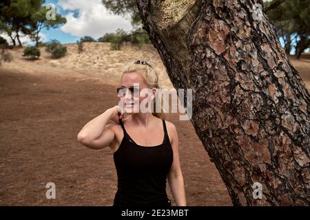 Glückliche junge Frau mit langen blonden Haaren berühren Sommer top Gurt und wegschauen, während in der Nähe von Baum im Park stehen In Madrid Stockfoto