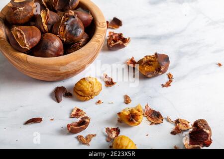Eine Holzschale aus hausgemachten, im Ofen gerösteten Kastanien mit Muscheln und Schalen auf der Marmorplatte verstreut. Ein traditioneller Winter-, Herbst-Snack mit Stockfoto