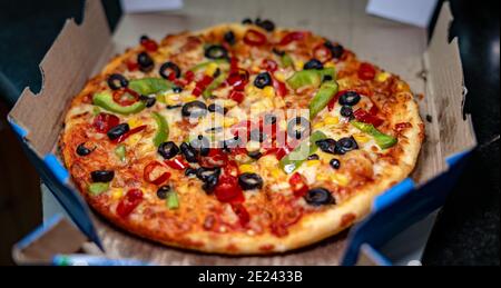 Eine vegetarische Pizza in der Pfanne mit schwarzen Oliven, grünen Paprika, rotem Paprika und goldenem Mais in einer Takeout-Box auf schwarzem Granit Stockfoto