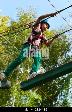 Ein Junge in einem Helm auf dem Kopf überwindet Hindernisse im Seilpark. Der Teenager bewegt sich auf Hängebalken mit einem Sicherheitsnetz. Stockfoto