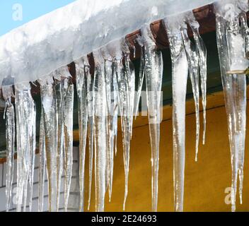 Eine Nahaufnahme von großen gefrorenen Eiszapfen von einem schneebedeckten Dach mit einer gebrochenen Regenrinne im Winter. Stockfoto