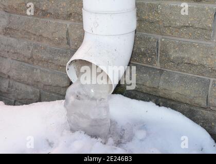 Eine Nahaufnahme eines Dachrinnenabwurfrohrs, ein Wasserauslauf mit gefrorenem Wasser, Eiszapfen in der Nähe des Hausfundaments im Winter. Stockfoto