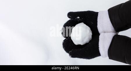 Weibliche Hände in schwarz gestrickten Fäustlingen halten einen Schneeball an Ein Schneehintergrund mit Kopierraum Stockfoto