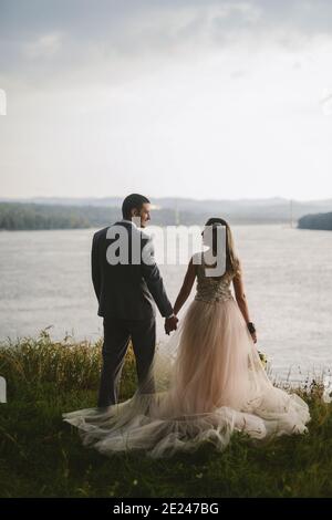 Gerade verheiratetes Paar, das in der Natur die Hände hält und einen schönen Blick auf den Fluss hat. Genießen Sie jeden Moment an ihrem besonderen Tag. Stockfoto