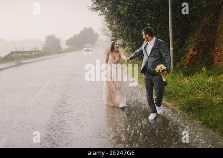 Gerade verheiratetes Paar, das Hände hält und auf Regen läuft. Spaziergang in nasser zeremonieller Kleidung auf der Fahrstraße. Lächeln und Spaß haben. Stockfoto