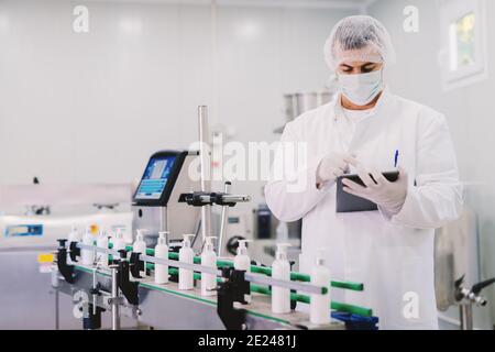 Bild von jungen Mann in steriler Kleidung mit Tablette. Steuerung der Produktionslinie im Werk. Flaschen mit kosmetischen Produkten auf Produktionslinie. Stockfoto