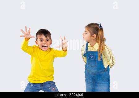 Der Junge macht gruselige Gesichter an der Kamera. Das Mädchen schaut ihn aufmerksam an. Weißer Hintergrund. Stockfoto