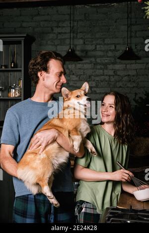 Glückliches kaukasisches Paar, das in der Küche mit ihrem Corgi-Hund kocht. Junge Familie, Frau und Mann essen zu Abend Stockfoto