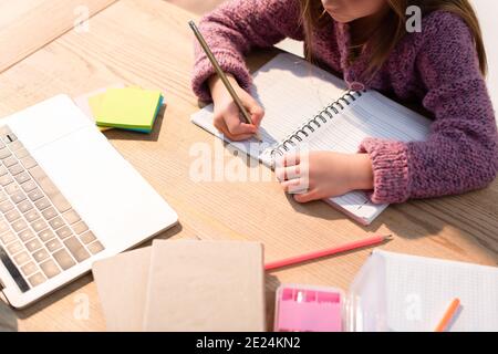 Beschnittene Ansicht des Mädchens, das im Notizbuch neben Haftnotizen, Büchern und Laptop auf dem Schreibtisch schreibt Stockfoto