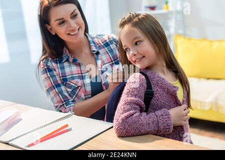 Glückliche Mutter, die zu Hause in der Nähe des Schreibtisches einen Rucksack auf die Tochter legt Auf unscharfem Hintergrund Stockfoto