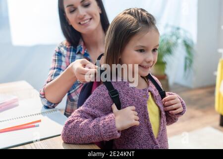 Lächelnde Mutter, die Rucksack auf Tochter zu Hause auf verschwommen Hintergrund Stockfoto