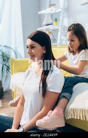 Tochter Flechten Haare von lächelnden Mutter, während sie auf der Couch sitzt Zu Hause auf unscharfem Hintergrund Stockfoto