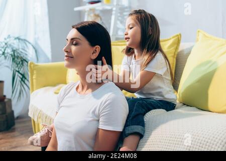 Tochter Flechten Haare lächelnde Mutter mit geschlossenen Augen während Sitzen auf Couch auf verschwommenem Hintergrund Stockfoto