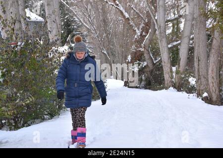 Warm gewickelte ältere Frau in Jacke, Stiefel und Hut zu Fuß auf einer verschneiten Landstraße. Szene nach dem Schneesturm namens Filomena in Spanien. Januar 2021. Stockfoto