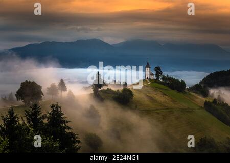 Jamnik, Slowenien - magischer nebliger Sommermorgen an der Kirche von Jamnik St.Primoz. Bei Sonnenaufgang. Der Nebel geht sanft hinter die kleine Kapelle mit golde Stockfoto