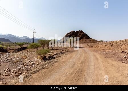 Ländliche Schotterstraße in Hatta Dorf im Hajar Gebirge in den Vereinigten Arabischen Emiraten mit kargem Land und trockenen Akazienbäumen. Stockfoto