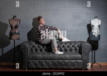 Ein junges Mädchen in legerer Kleidung sitzt auf der Rückseite eines Ledersofas gegen eine graue Ziegelwand. Das Model trägt ein kariertes Hemd, Jeans und weiße Sneakers Stockfoto