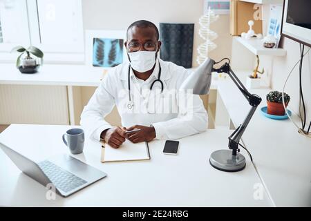 Arzt in einer Maske posiert für Kamera an seinem Schreibtisch Stockfoto