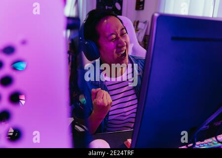 Junge asiatische männliche Gamer gewann und Sieg das siegreiche Spiel mit Headset und pc-Computer. Überglücklich live Streamer Streaming Video-Spiel in wenig Licht Raum. Begeisterter Gaming-Influencer lächelt. Mit Kopierbereich Stockfoto