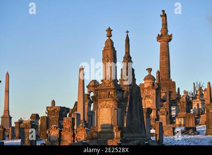 Nahaufnahme einer Gruppe von Grabsteinen und Denkmälern, die an einem verschneiten Wintertag in der Glasgow Necropolis durcheinander kamen. John Knox Denkmal an der Spitze. Stockfoto