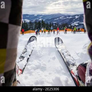 Nahaufnahme eines Mannes auf Skiern, der auf weißem Tiefschnee im Skigebiet steht. Alpine Skier an Skischuhen mit Skibindungen befestigt und Gruppe von Menschen mit Bergen auf verschwommenem Hintergrund. Konzept des Skifahrens Stockfoto