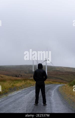 Eine Kapuzenfigur, die auf einer Feldbahn steht. Blick auf Windenergieanlagen. Auf einer düsteren Bergstrecke. Wales. VEREINIGTES KÖNIGREICH. Stockfoto