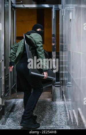 Maskierter Dieb, der nachts mit gestohlenem Fernseher aus dem Gebäude entkommen ist Stockfoto