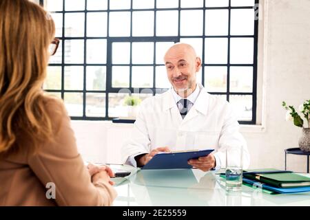 Schuss eines männlichen Arztes mittleren Alters, der mit seinem Patienten spricht, während er am Büroschreibtisch im Arztraum sitzt. Stockfoto