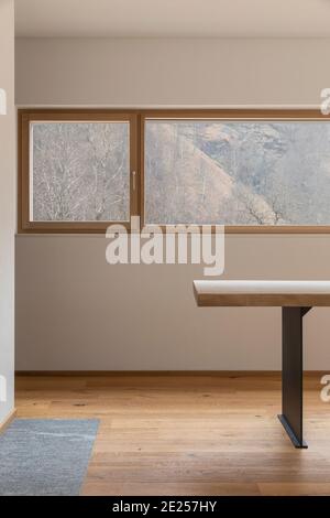 Modernes Interieur mit weißen Wänden und Parkettböden. Detail von Holztisch mit Design-Lampe und Fenster zum Wald der Schweizer Alpen. Niemand Stockfoto