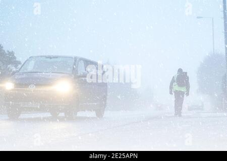 Rückansicht eines männlichen Fußgängers, der an einem Morgen des 2021. Januar auf vereister Fahrbahn läuft, während starker Schnee fällt und der Verkehr in eisigen Nebel vorbeigeht. Stockfoto