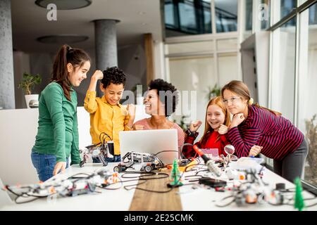 Gruppe von glücklichen Kindern mit ihrer afroamerikanischen weiblichen Wissenschaft Lehrer mit Laptop Programmierung von elektrischen Spielzeugen und Robotern bei Robotics Schulungsraum Stockfoto