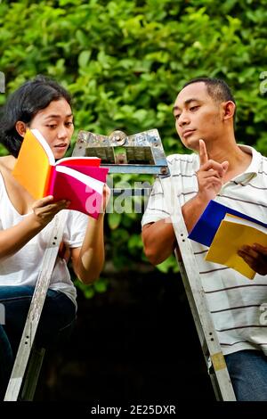 Südostasiatischer junger Mann und Frau, die auf einer Leiter sitzen, zusammen studieren und über Bücher diskutieren Stockfoto