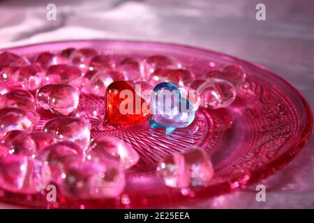Ein rotes und ein blaues Glas Herz umgeben von rosa Herzen auf einem rosa Glasplatte mit Ornamentmuster. Stockfoto