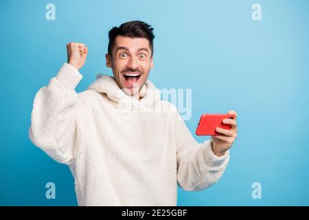 Foto-Porträt von schockierten triumphalen Mann hält Telefon in einem Hand vertikal tragen Wolle Hoodie isoliert auf pastellblau gefärbt Hintergrund Stockfoto