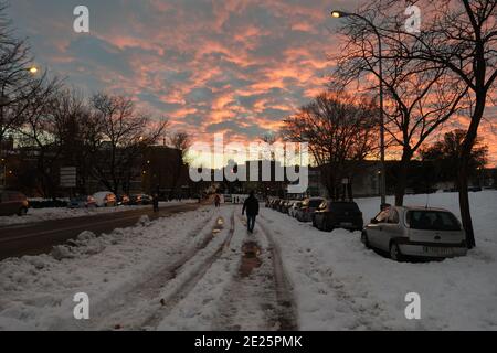 Madrid, Spanien. 10. Januar 2021: Sonnenuntergang in einer verschneiten Landschaft auf einer Straße, mit Menschen, Autos und Wolken während Filomena Schneesturm. Stockfoto